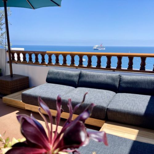 - Relax on the plush outdoor sectional under a large umbrella with uninterrupted sea views
- Tropical plants and a distant cruise ship add vibrant charm to this sunny deck
- Perfect for unwinding with cocktails or catching golden sunsets